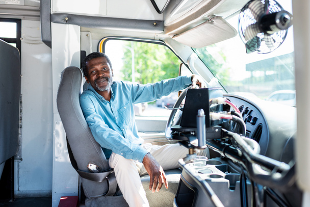 uśmiechający się dojrzałe african american bus sterownika patrząc na kamery podczas siedzenia wewnątrz autobusu - Zdjęcie, obraz
