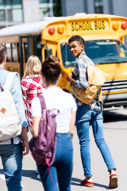 vista posteriore di bello teen africano americano scolaro a piedi a scuolabus con i suoi compagni di classe e tornando alla fotocamera
 - Foto, immagini