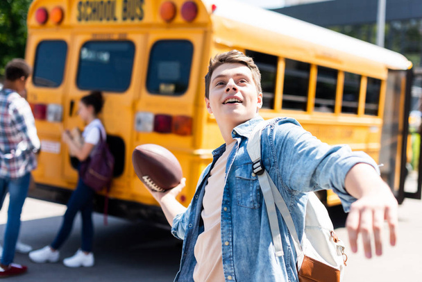 gülümseyen genç öğrenci okul otobüsü önünde Amerikan futbolu topu atma - Fotoğraf, Görsel
