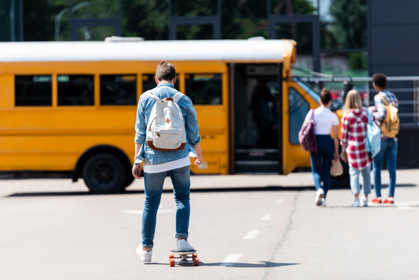 rear view of schoolboy riding skateboard by parking to his classmates - Foto, Imagen