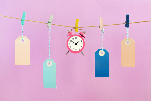 On the ropes on the clothespins are hung empty blue and pink labels. In the center is a small alarm clock. Concept, time to buy. Beautiful layout on a pink background. - Photo, Image
