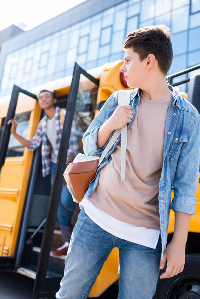 vista inferior de adolescente colegial caminando en frente de autobús escolar y volver a su compañero de clase
 - Foto, Imagen