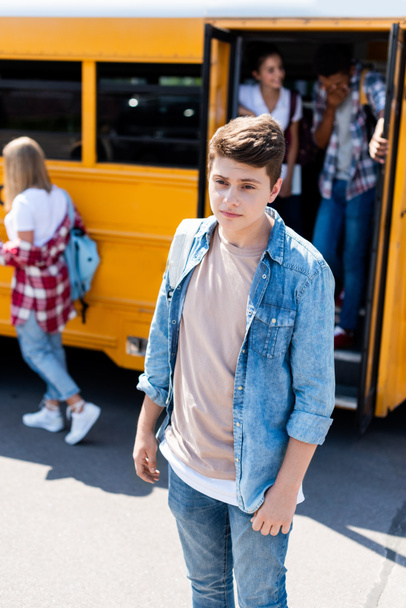 depressed teenager schoolboy standing in front of school bus with classmates - Foto, afbeelding