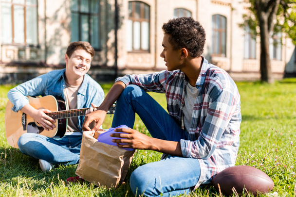 happy teenagers sitting on grass and playing acoustic guitar during lunch - Foto, Imagen