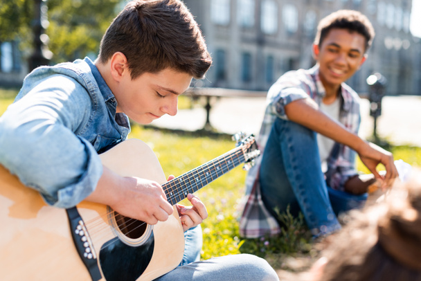 happy teenagers sitting on grass and playing acoustic guitar - Fotó, kép