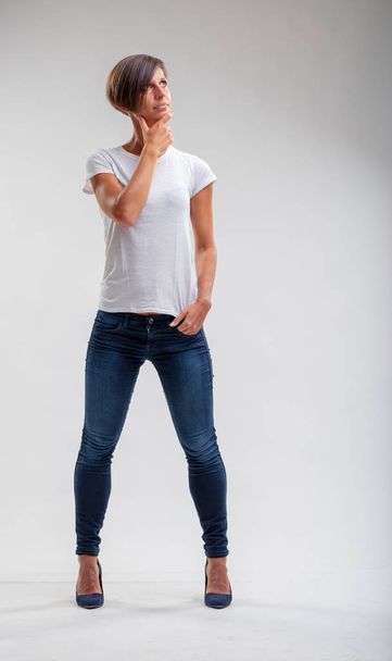 Mujer joven musculosa en pantalones vaqueros azules casuales de pie con la mano en la barbilla mirando hacia arriba a un lado sobre blanco con espacio para copiar
 - Foto, Imagen