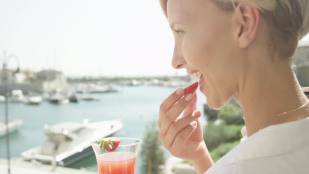 Slow Motion Close Up Dof: Wesoła dorosła kaukaska kobieta na wakacjach jedząca słodkie truskawki i pijąca koktajle na hotelowym tarasie, ciesząca się widokiem na zatokę żaglową w niesamowity słoneczny letni dzień - Materiał filmowy, wideo