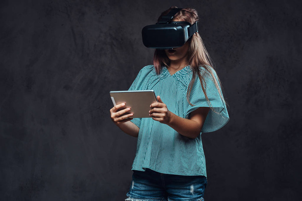Νεαρό κορίτσι με χρήση εικονικής πραγματικότητας γυαλιά και κατέχει έναν υπολογιστή tablet. Απομονωμένα σε σκούρο φόντο με υφή. - Φωτογραφία, εικόνα