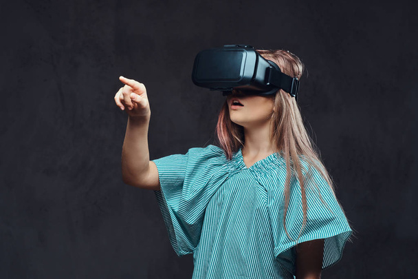 Young girl dressed casual using virtual reality glasses. Isolated on a dark textured background. - Фото, изображение