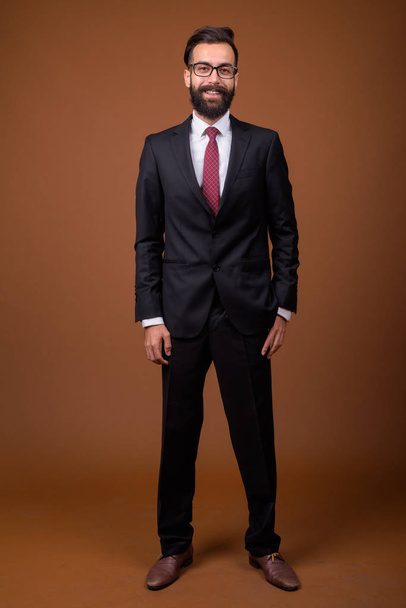 Studio shot of young handsome bearded Persian businessman wearing eyeglasses against brown background - Foto, immagini