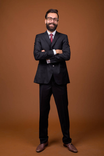 Studio shot of young handsome bearded Persian businessman wearing eyeglasses against brown background - Foto, immagini