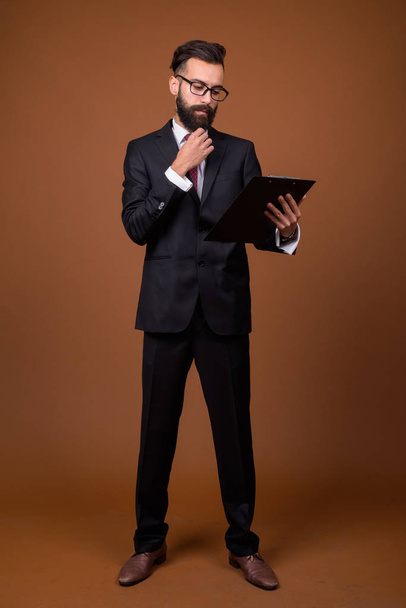 Studio shot of young handsome bearded Persian businessman wearing eyeglasses against brown background - 写真・画像