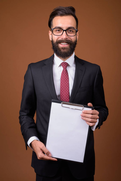 Studio shot of young handsome bearded Persian businessman wearing eyeglasses against brown background - Foto, Imagen