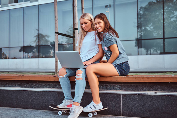 Dos hermosas chicas hipster sentadas en el banco con una computadora portátil en el fondo del rascacielos
. - Foto, Imagen