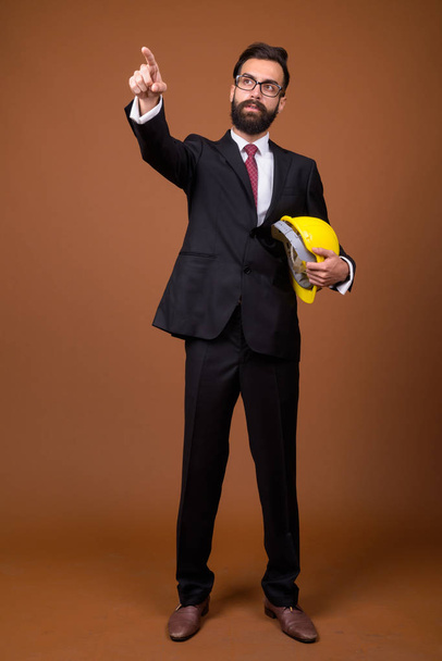 Studio shot of young handsome bearded Persian businessman with hardhat wearing eyeglasses against brown background - Photo, image