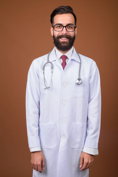 Studioaufnahme eines jungen gutaussehenden bärtigen persischen Arztes vor braunem Hintergrund - Foto, Bild