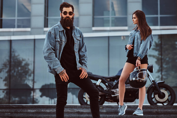 Casal hipster atraente - macho brutal barbudo em óculos de sol vestidos com uma jaqueta de jeans e sua jovem menina sensual em pé em passos contra arranha-céus
. - Foto, Imagem