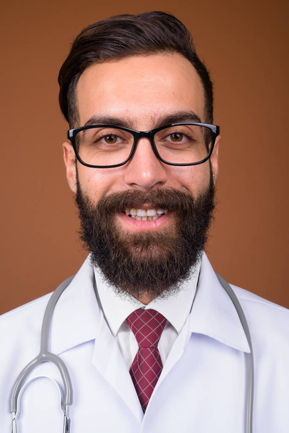 Studio shot of young handsome bearded Persian man doctor against brown background - Photo, Image