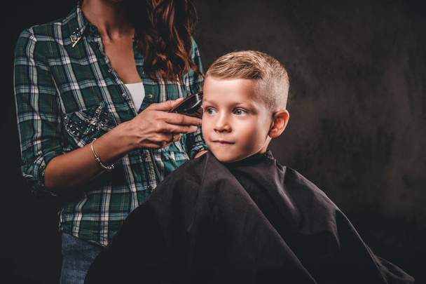子供美容師トリマーには、暗い背景に小さな男の子を切っています。かわいい幼児少年取得散髪.  - 写真・画像