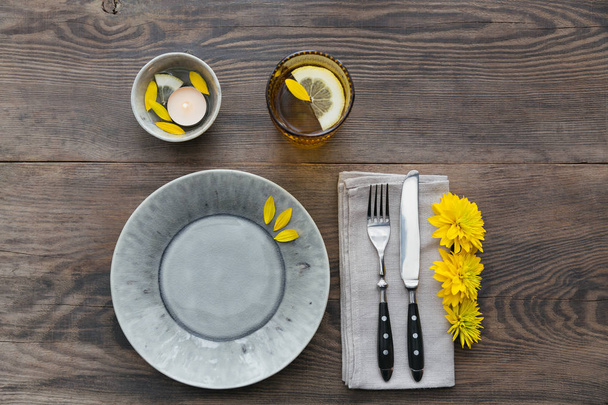 Rustic table setting with linen napkin, cutlery, ceramic plates, yellow glasses and yellow flowers on dark wooden table. Holiday table decoration. Romantic dinner. - Фото, изображение