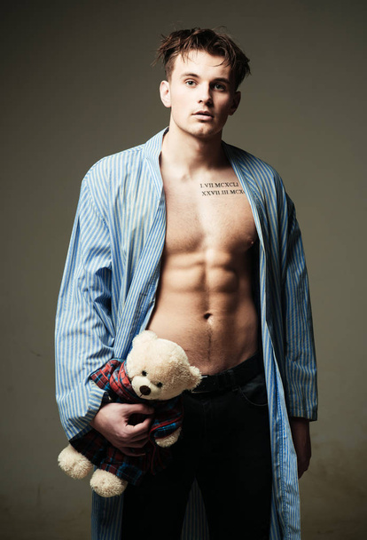 Guy in bathrobe with romantic gift. Man on calm face, muscular figure, holds soft toy plush bear in hand. Man with muscular torso, six packs, looks attractive, dark background. Romantic macho concept. - Foto, Imagen