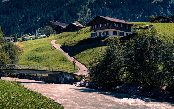 Malowniczym alpejskim sceny z rzeką, niektóre typowe drewniane domki i zielonym turystą retro pociąg, zejście z góry. Grindelwald, regionie Oberland Berneński, Szwajcaria - Zdjęcie, obraz