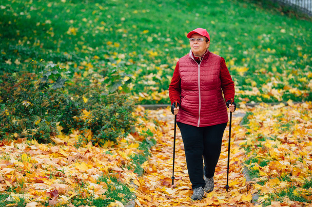 Senior woman standing with nordic walking poles in colorful autumn park near river. Healthy life concept. Mature woman resting after exercise outdoors. - Fotó, kép