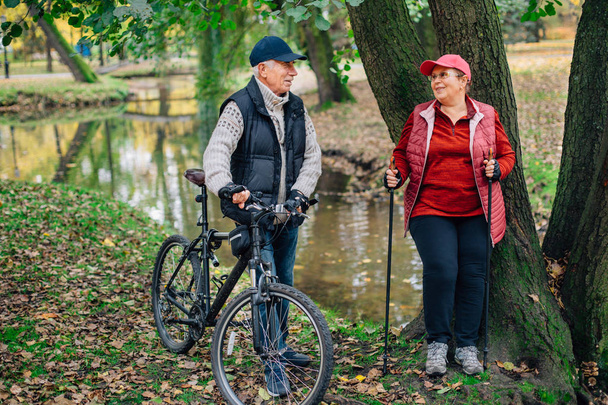 Bella coppia di anziani sport in piedi in colorato parco autunnale vicino al fiume. Donna matura con bastoni da nordic walking e vecchio con bicicletta che riposa all'aperto
. - Foto, immagini