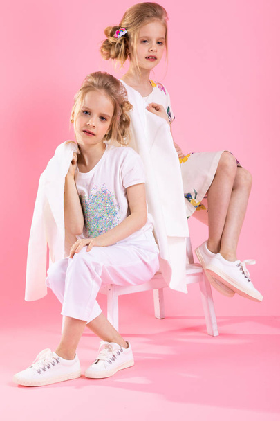 Twins girls in bright clothes posing near the stairs with two steps on a pink background. - Photo, Image