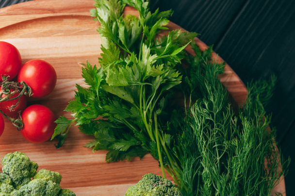 On a dark wooden table, on a round wooden board fresh green broccoli, parsley, dill and cherry tomatoes for your health. Recipe. Ingredients. Dietary food. Place under the text. View from above - Photo, Image
