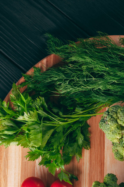 On a dark wooden table, on a round wooden board fresh green broccoli, parsley, dill and cherry tomatoes for your health. Recipe. Ingredients. Dietary food. Place under the text. View from above - Foto, Imagen