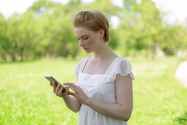 Cute Kobieta czyta przyjemne wiadomości tekstowych na telefon komórkowy siedząc w parku w ciepłym dniu wiosny, wspaniała kobieta słucha muzyki i wyszukiwanie informacji na telefon komórkowy - Zdjęcie, obraz