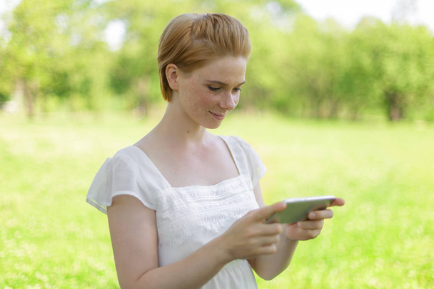 Χαριτωμένο γυναίκα διαβάζει ευχάριστο μήνυμα κειμένου στο κινητό τηλέφωνο ενώ κάθονται στο πάρκο σε ζεστή ημέρα άνοιξη, πανέμορφο θηλυκό είναι ακούγοντας μουσική και αναζητώντας πληροφορίες στο κινητό τηλέφωνο - Φωτογραφία, εικόνα
