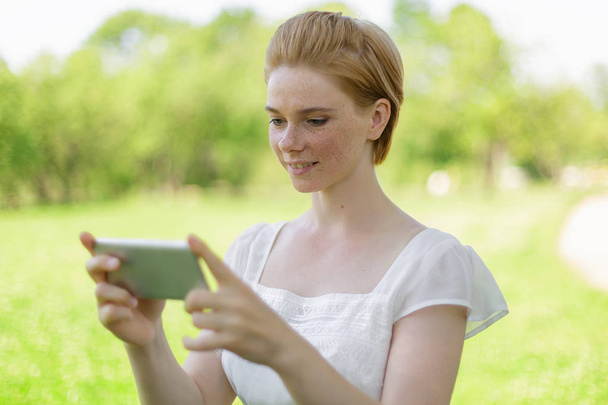 かわいい女性は暖かい春の日に公園に座っている間、携帯電話で楽しいテキストメッセージを読んでいる、ゴージャスな女性は、音楽を聴いて、携帯電話で情報を検索しています - 写真・画像