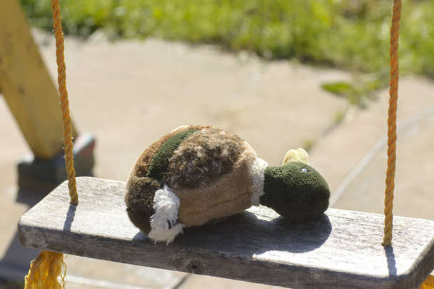 Мягкая детская игрушка (плюшевая утка) лежит на детской скамейке для верховой езды, привязанной желтыми веревками. Концепция летнего отдыха для детей
. - Фото, изображение