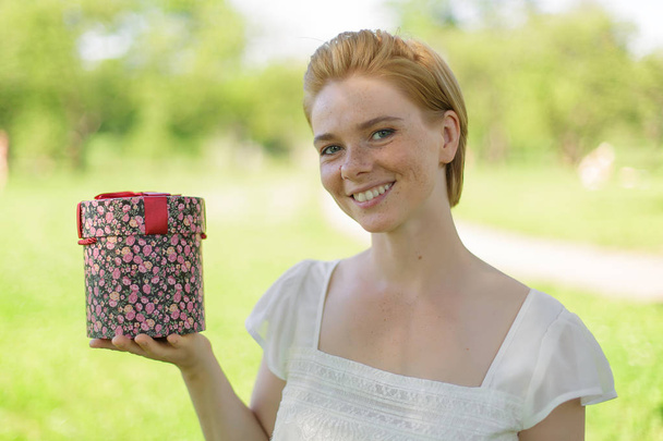 Tämä on sinulle. Houkutteleva nuori nainen, jolla on lahjapakkaus käsissään seisoo kesän vihreää puistoa vastaan
 - Valokuva, kuva