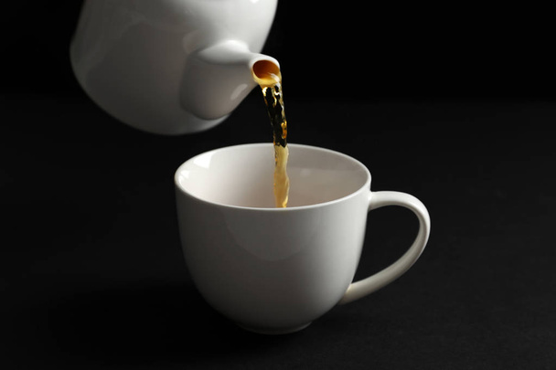 Pouring hot tea into white porcelain cup on dark background - Foto, afbeelding