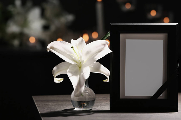 Funeral photo frame with black ribbon and lily on table in dark room - Foto, Imagem