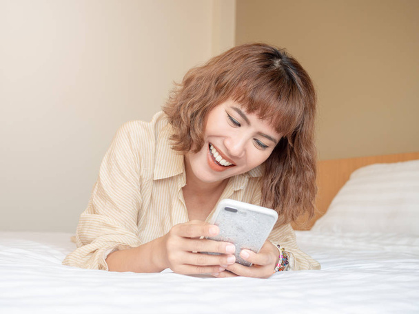 Ευτυχής όμορφη γυναίκα της Ασίας γελώντας ξαπλωμένη στο κρεβάτι white χρησιμοποιώντας κινητό τηλέφωνο κοντά μαλλιά. - Φωτογραφία, εικόνα