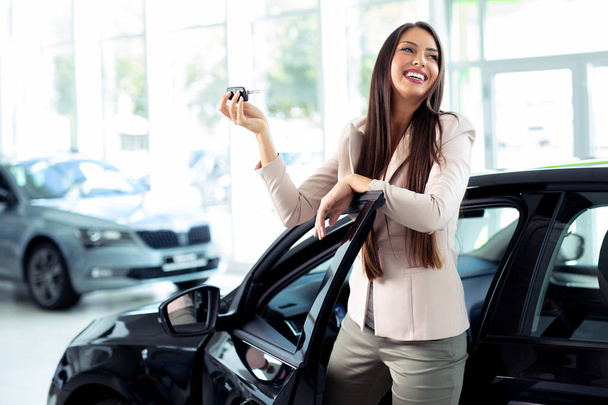 Happy woman holding keys to her new car at the dealership - Fotoğraf, Görsel