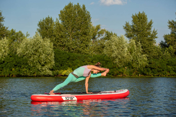 Mujer joven haciendo yoga en sup board con remo. Postura de yoga, vista lateral - concepto de armonía con la naturaleza
. - Foto, Imagen