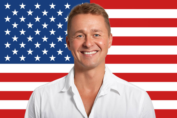 Happy European male with pleasant smile, dressed in white shirt, rejoices positive moments in life, stands against American flag in studio, expresses positiveness and happiness. Masculinity concept - Photo, image