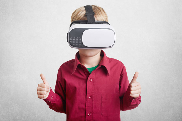 Pleased small boy enjoys virtual reality, wears VR headset or 3D glasses, shows ok sign as being satisfied with its effect, plays video games online isolated on white background. Innovative technology - Фото, зображення