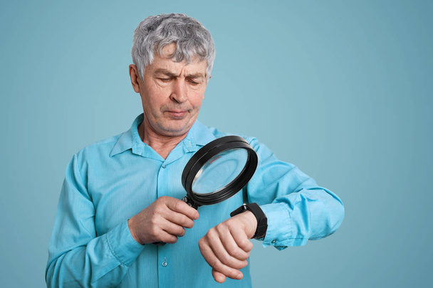Technology, health care and pension concept. Senior mature male looks at smartwatch through lens, checks puls, controls his health, dressed in formal shirt, isolated over blue studio background - Foto, Imagem