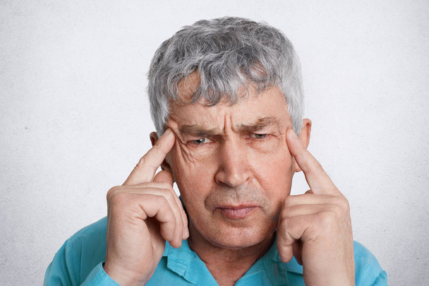 Close up portrait of wrinkled grey haired mature man keeps fingers on temples tries to find solution in mind or remember something, wears formal clothing, isolated over white studio background - Photo, Image