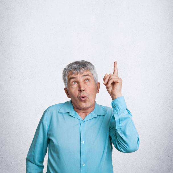 Indoor vertical shot of amazed elderly male indicates upwards as notices something surprising, dressed in elegant shirt, poses against white background with copy space for your advertisement - Фото, зображення