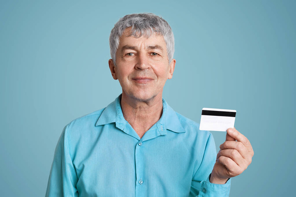 Pleased mature male with cheerful expression in formal shirt, holds credit plastic card, happy to get pension on it, isolated over blue background. Content boss in formal clothes poses in studio - Valokuva, kuva