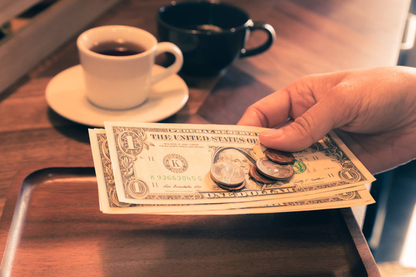 Kobieta ręka trzyma nas Dolar banknotów i monet w drewniana Taca na stole kawiarni, dwie filiżanki herbaty na tle, Styl vintage. Pojęcia dotyczące płatności, opłaty za obsługę, bill, sprawdzanie, porady. - Zdjęcie, obraz