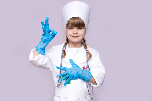 Sonriente niña médico en uniforme médico y guantes azules muestran símbolo ok
 - Foto, Imagen