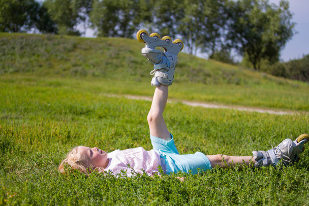 leisure, childhood, outdoor games and sport concept - Cute little blonde the little girl lies on the grass in roller skates - Photo, Image